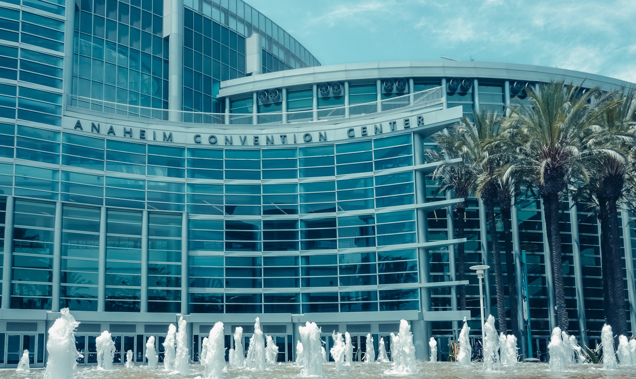 View of the Anaheim Convention Center on a sunny day, with palm trees and blue sky seen from across the main plaza and fountain.